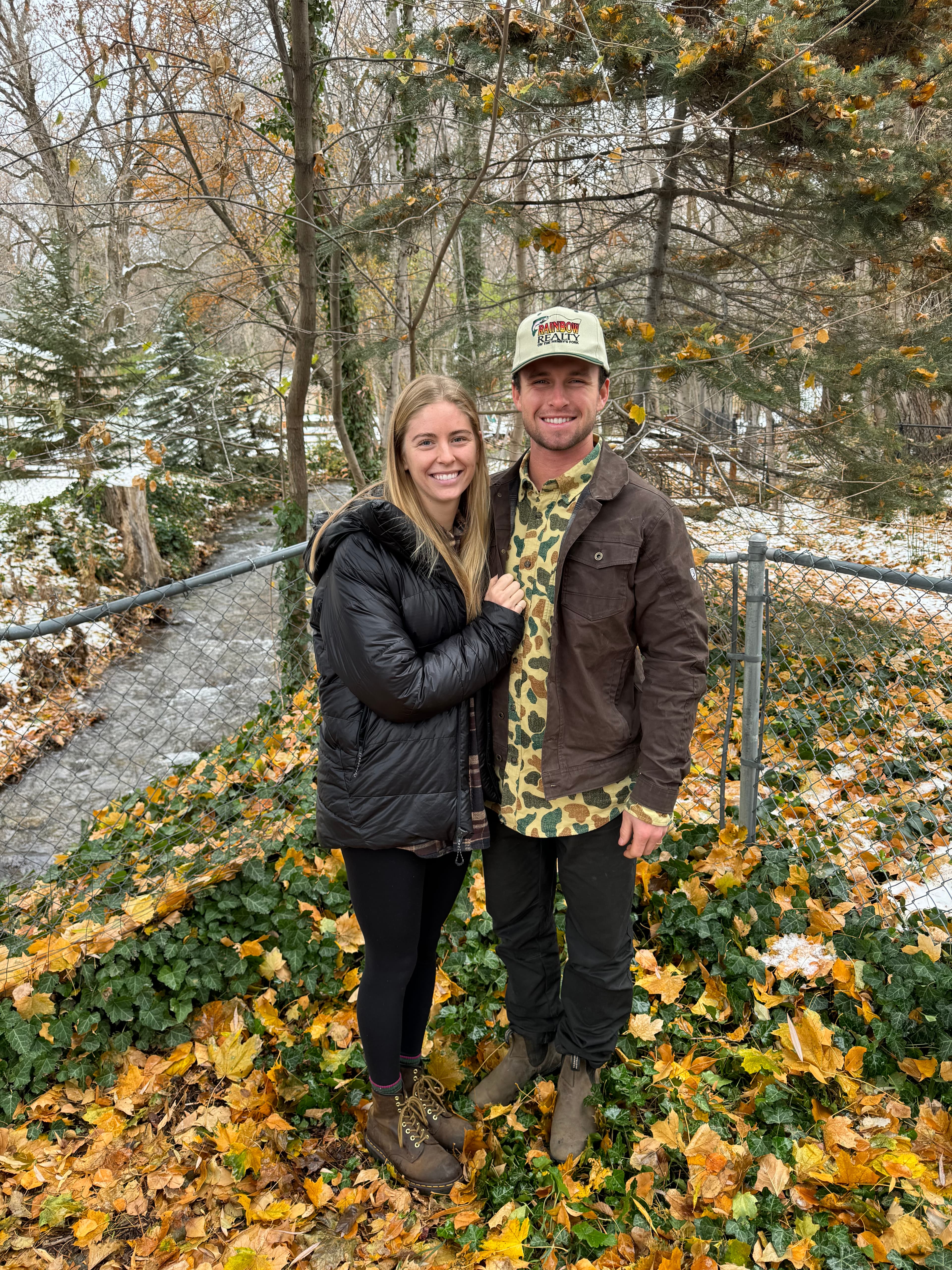 A smiling man and woman pose together outdoors amidst fallen yellow autumn leaves.