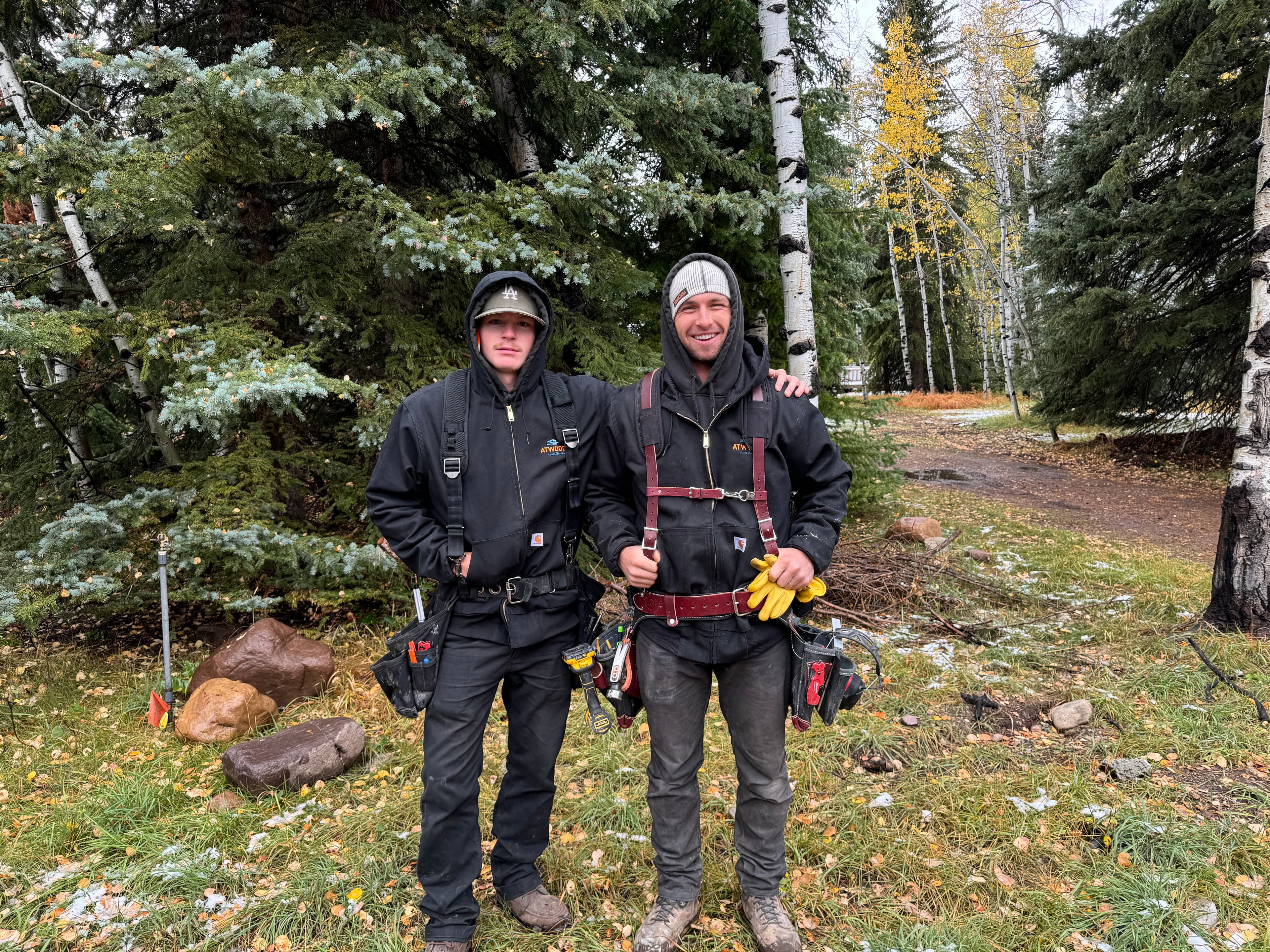 Two men in black workwear and tool belts stand together in a wooded landscape.